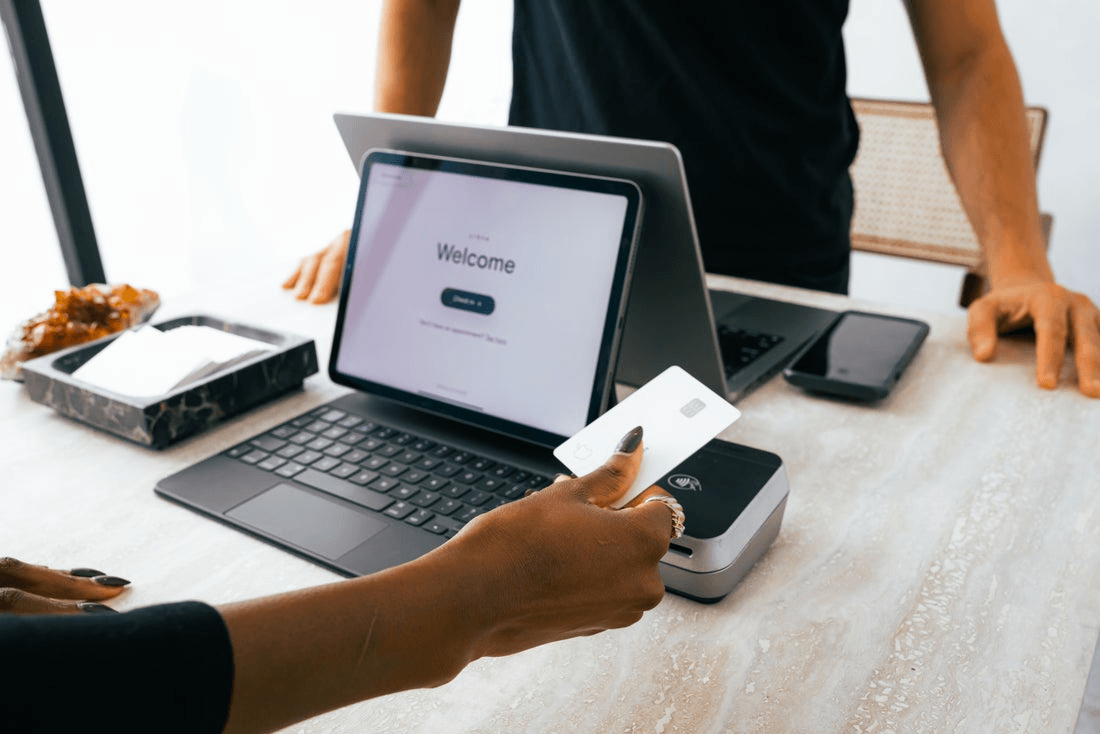 The front desk of a salon. Behind the counter, an employee stands with a laptop. The client faces a tablet and holds out a credit card.