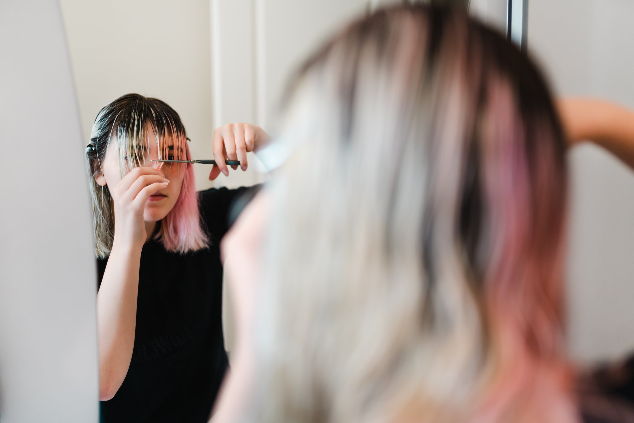 A young woman with bleached and pink hair cuts her own bangs in front of a mirror.