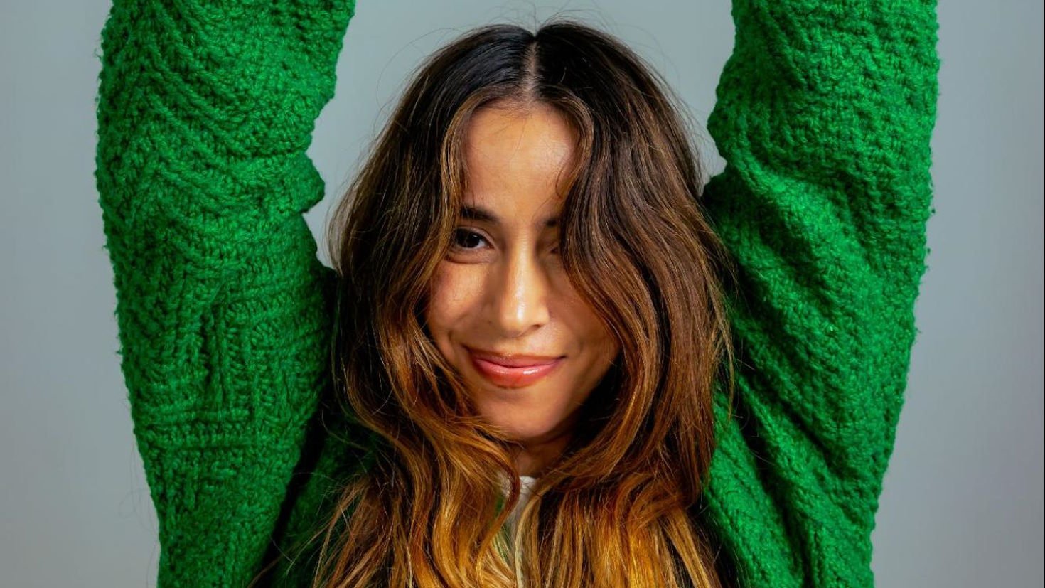 Colorist Jess Gonzalez smiles while raising both arms in the air and wearing a vibrant green sweater.