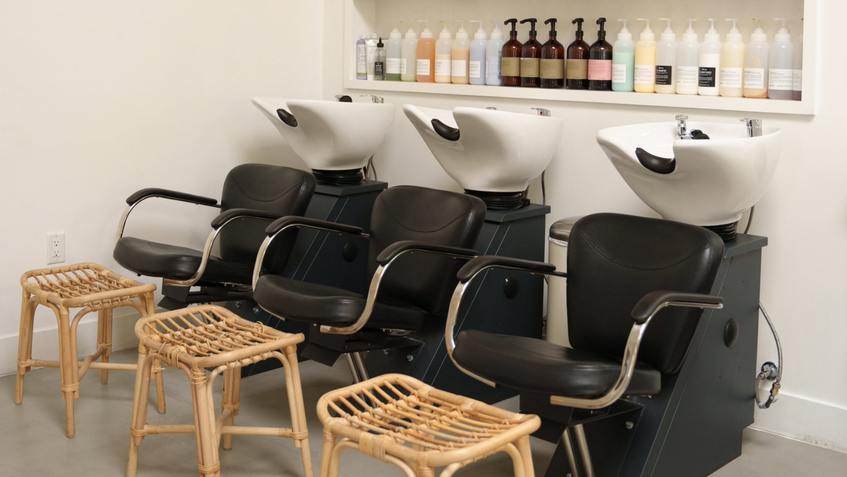 Salon hair washing station with 3 sinks, chairs, and stools in front of a wall with a shelf of hair products.