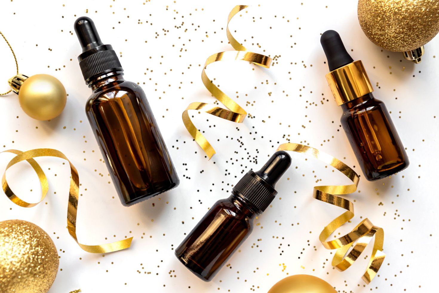 Tinctures of cosmetic products are scattered around a table. Interspersed between are golden ornaments, confetti, and ribbons.