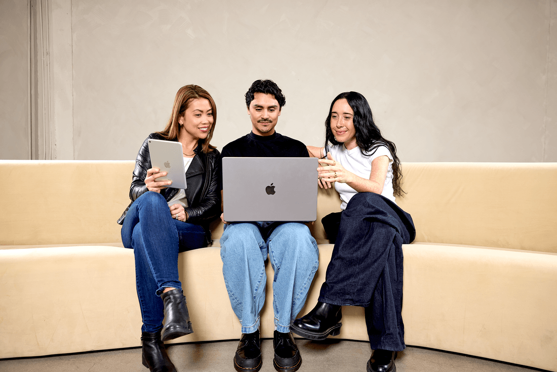 Two women and a man sit on a beige couch while looking at a digital tablet and a laptop.