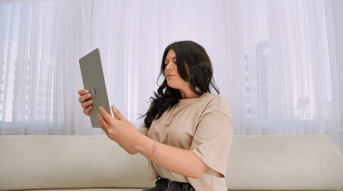 A medspa owner sits on a couch looking at her business reports on an iPad.