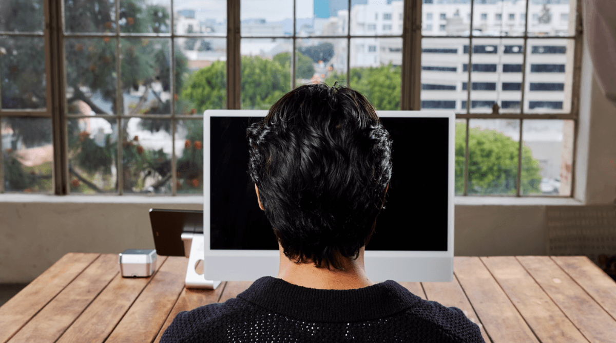 A man sits at a wooden desk and looks at a computer screen, a digital tablet, and a payment terminal.