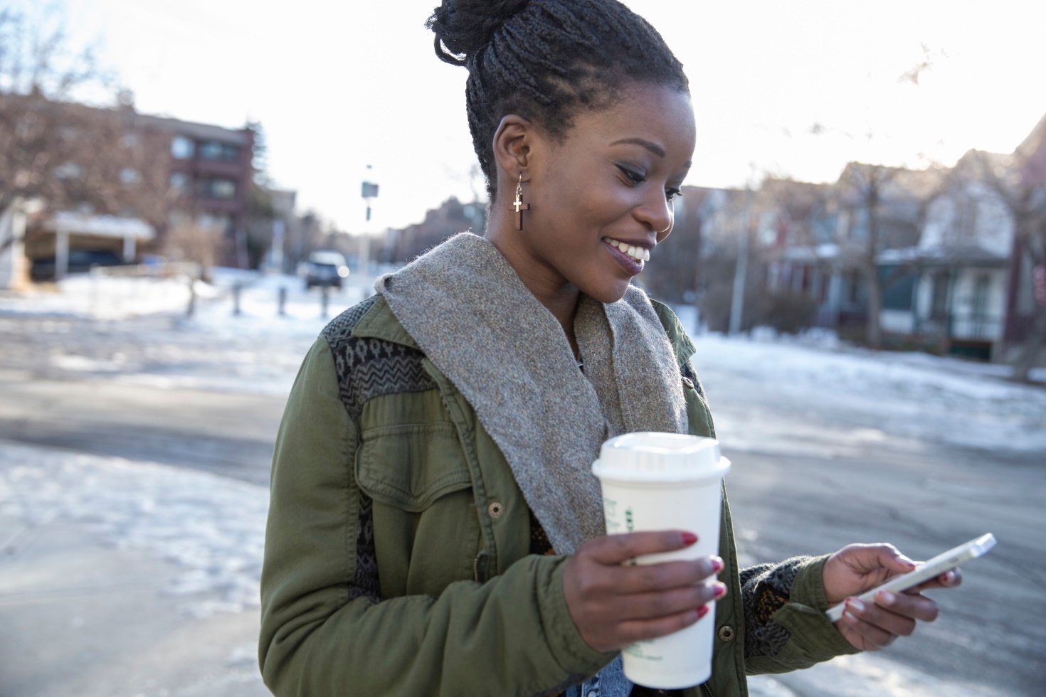 A woman holds coffee and checks her phone on a snow-covered street corner.