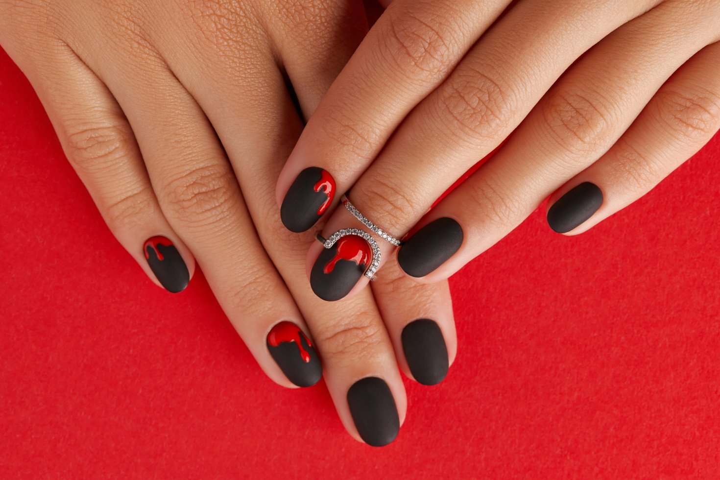 A pair of hands on a plain red background show off matte black fingernails with blood-crimson drips painted at the top.