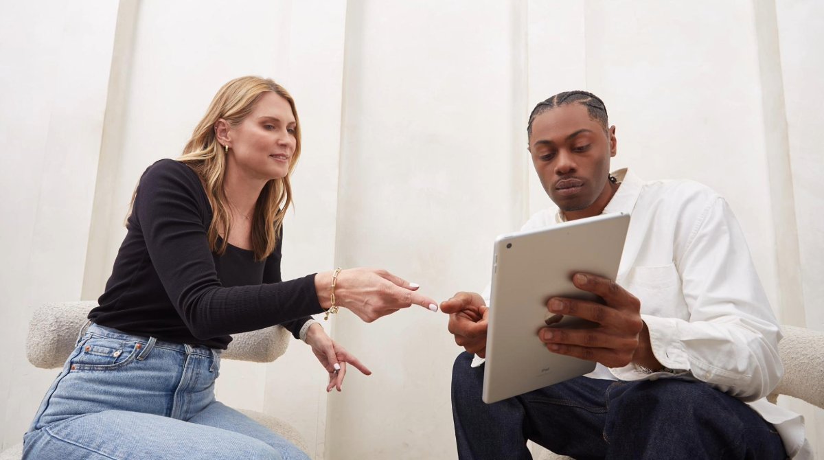 A woman and a man are sitting in chairs next to each other looking at a tablet.