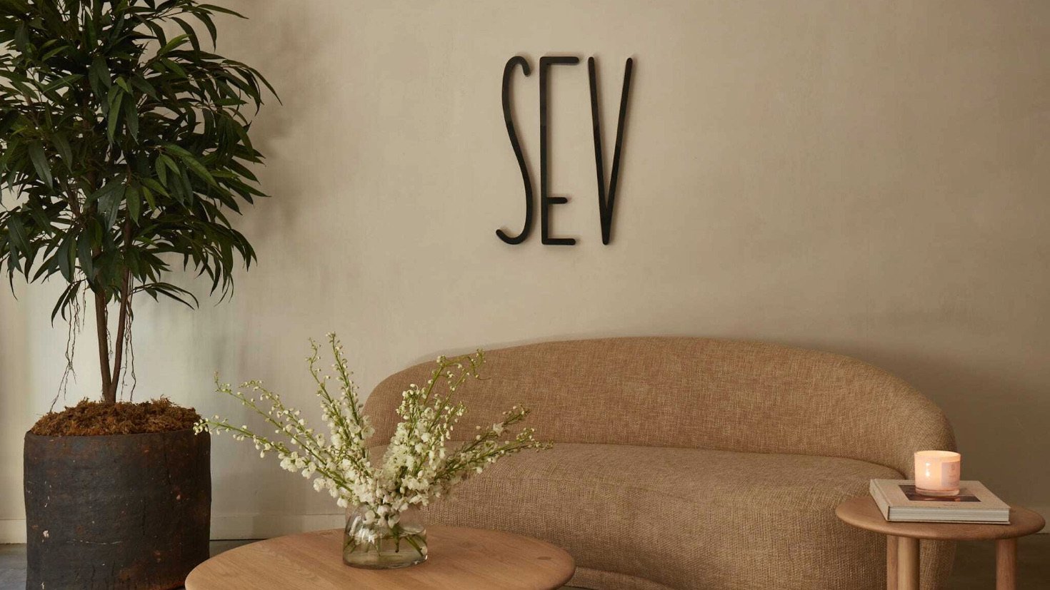 SEV Laser lobby with a tan couch, coffee table, and side table with a tall indoor plant shown with a sign on the wall displaying their name, "SEV"
