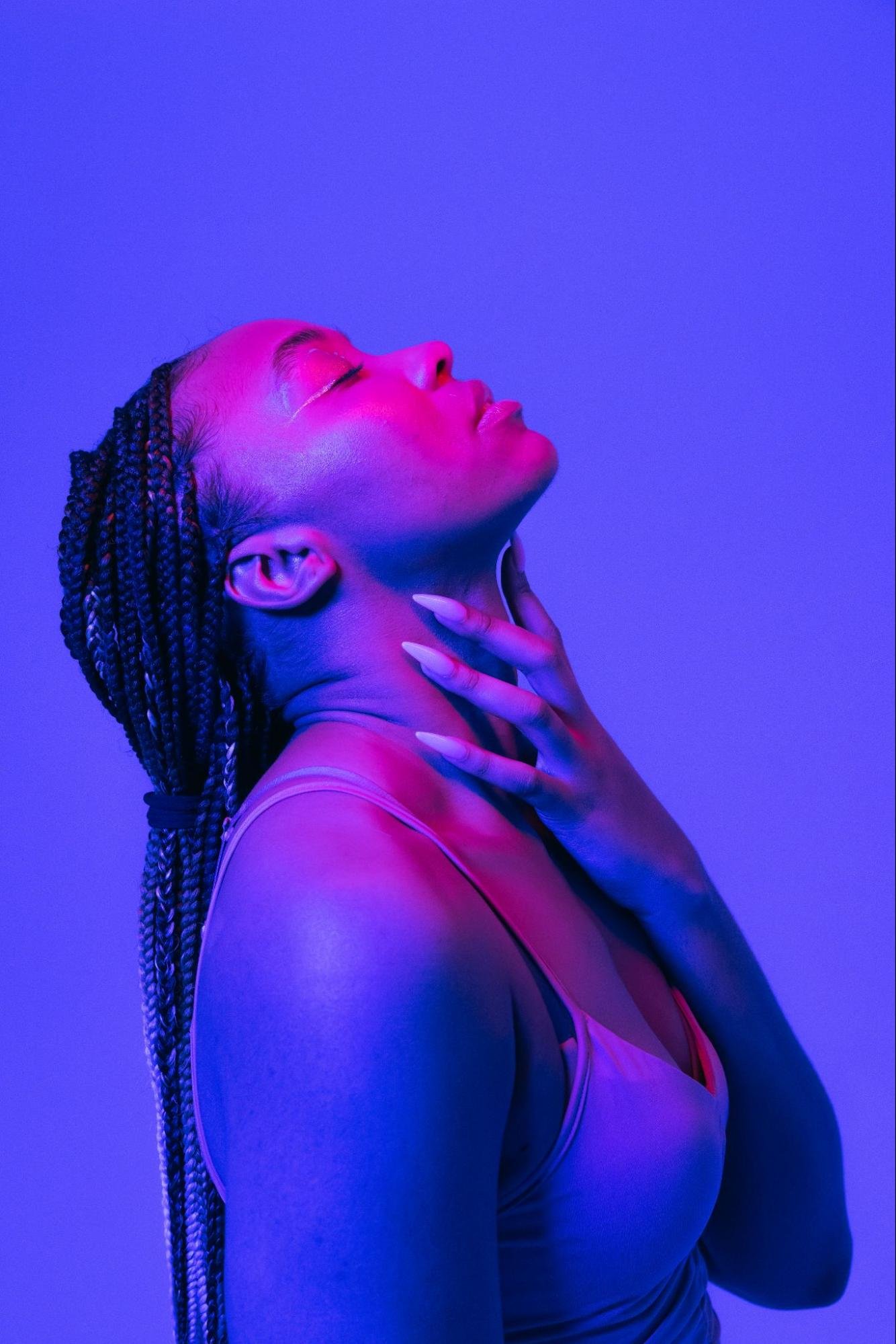 A person with long, braided hair and a white tank top leans their head back with their eyes closed. They are seen from the side and are lit in purples and pinks.