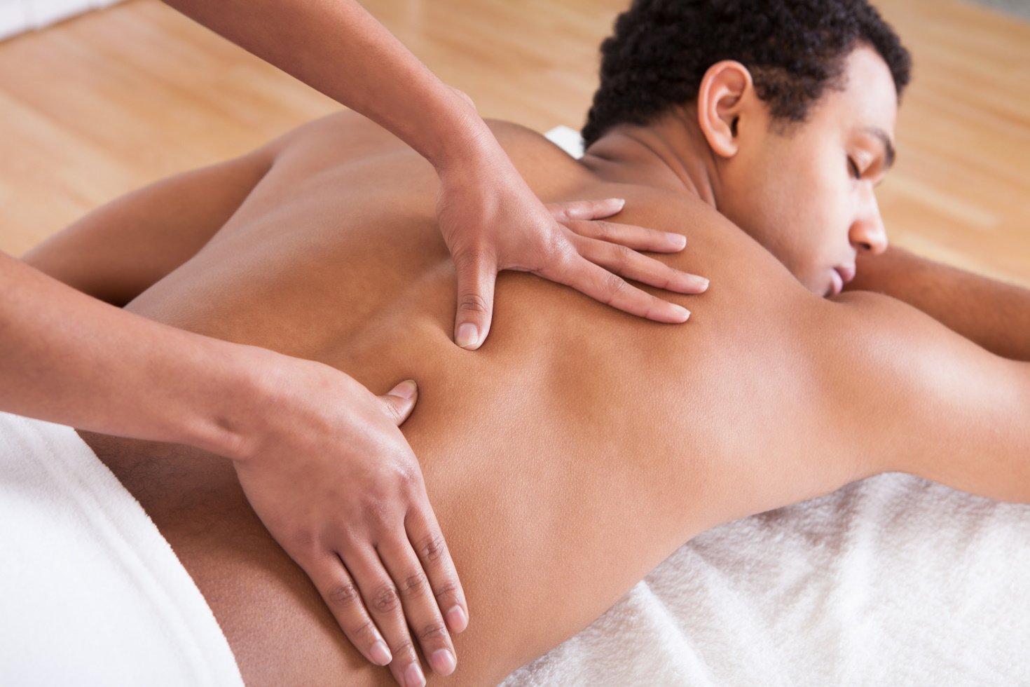 A man lies on a massage table with his eyes closed and a towel covering him from the waist down while two hands massage his back.