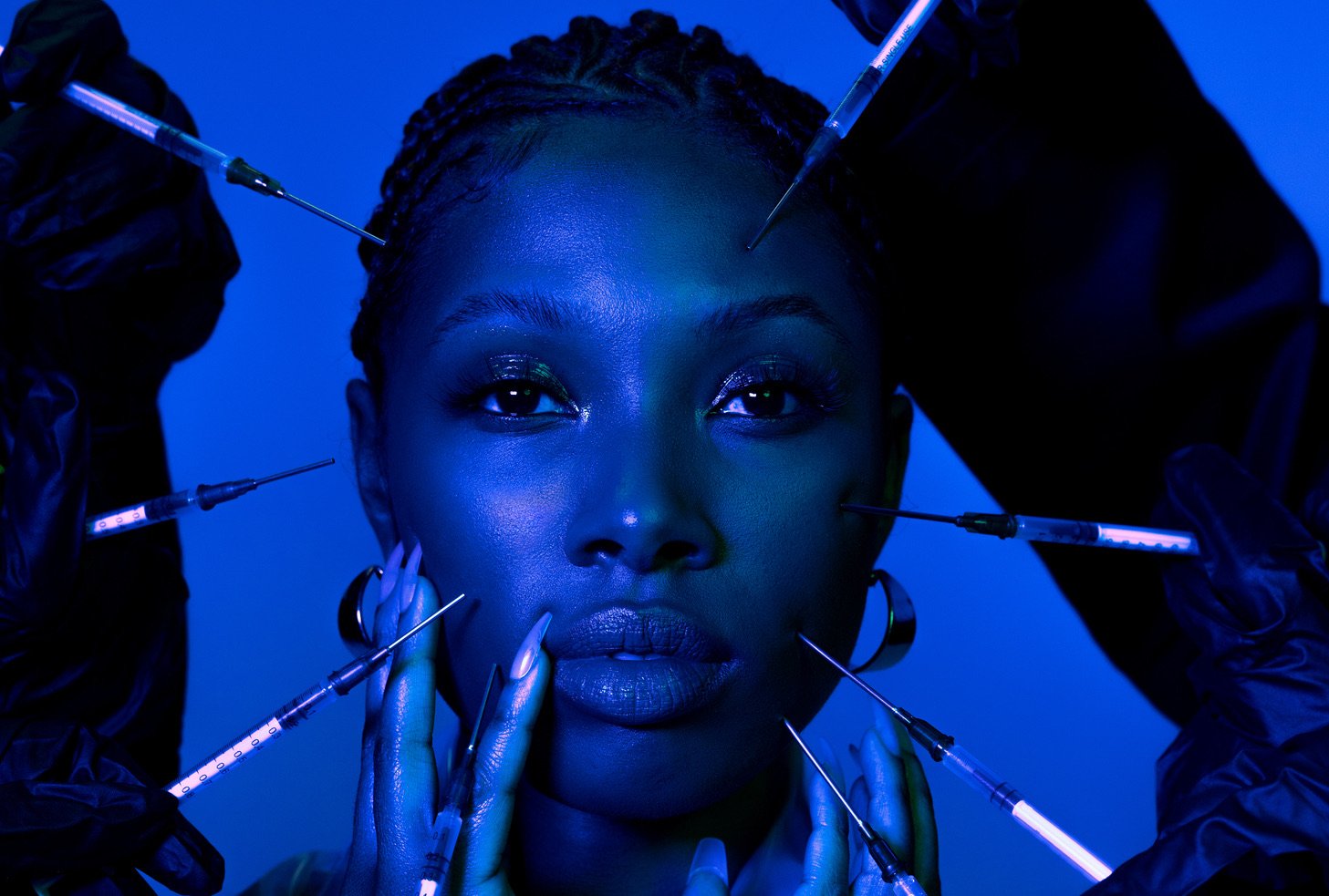A blue-tinted image of a medspa client with long nails, braids, and a transparent jacket. A set of eight syringes is arrayed in front of their face.