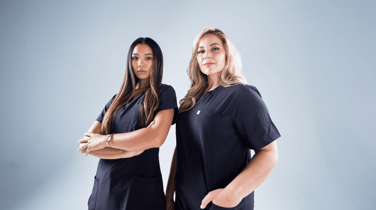 Two women in medical scrubs pose against a gray background.